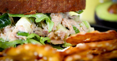 Toasted whole wheat toast, with tuna salad, avocado sliced and arugula. Avocado half in the background and thin pretzels in the foreground.