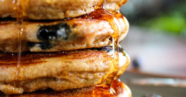Whole Wheat Blueberry Pancake stack, with a pad of butter, syrup, and fresh blueberries on a cyan plate, wood plank table and fork. 