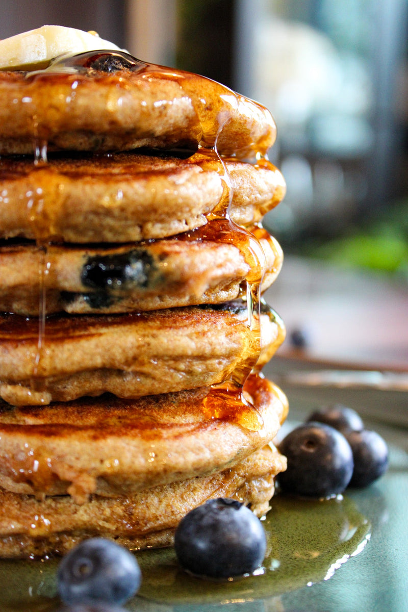 Whole Wheat Blueberry Pancake stack, with a pad of butter, syrup, and fresh blueberries on a cyan plate, wood plank table and fork. 