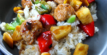 Close-up of sweet and sour chicken meatballs served over white rice, with caramelized red and green bell peppers and golden pineapple chunks coated in glossy sauce.